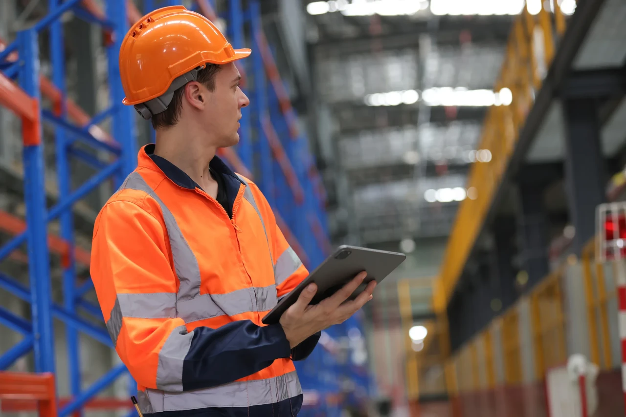 Factory or warehouse worker in safety vest reviewing data on a tablet, showing how computerized maintenance management software support daily operations